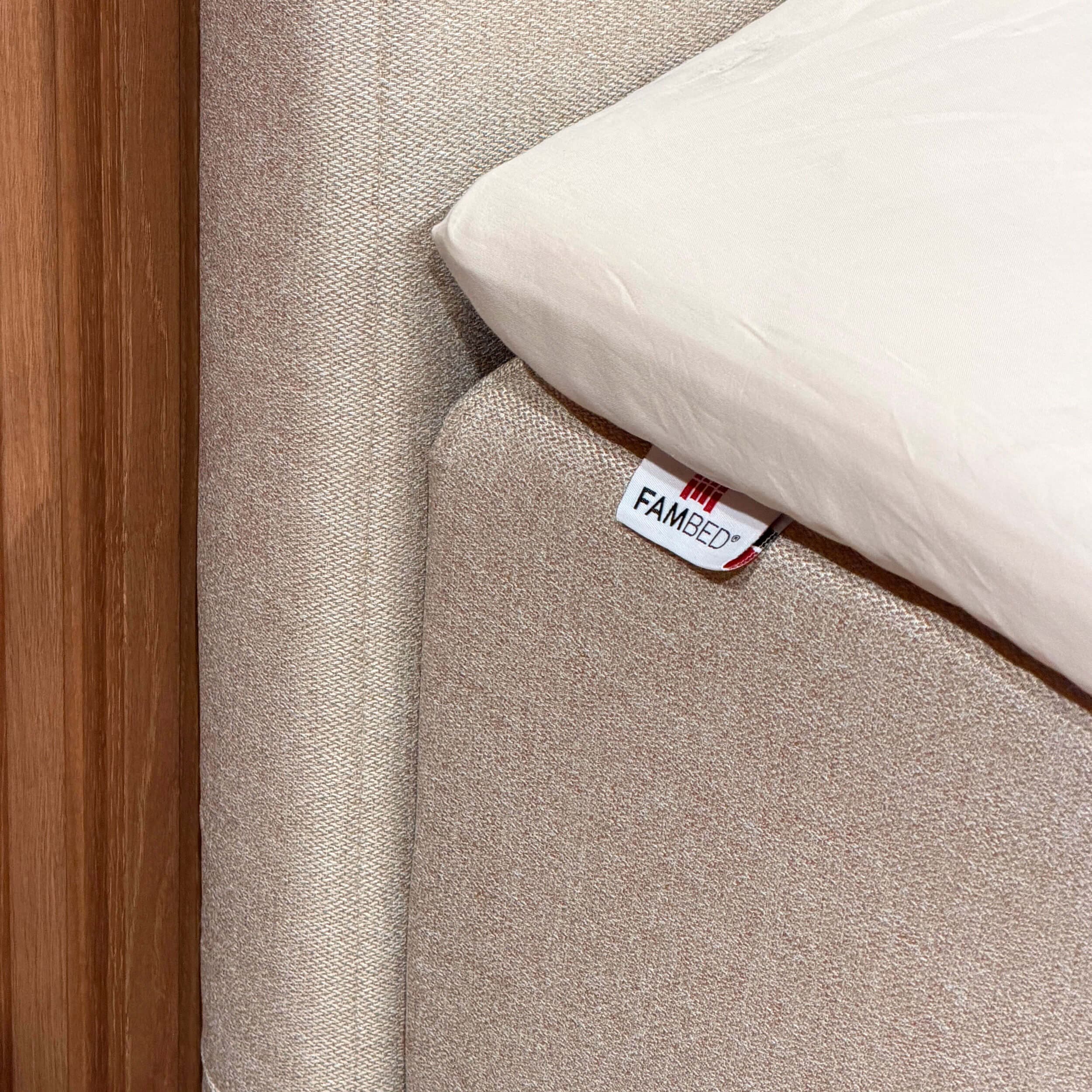 Close-up van de beige hoes rond de hoek van het bed, met een FAMBED-label en een wit laken erop