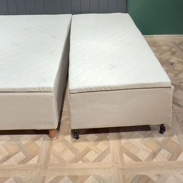 Twee volledig gemonteerde bedmodules staan naast elkaar op een lichte houten vloer, met een smalle kier ertussen. Elke module heeft een eigen beige bekleding, gewatteerde matras en poten.