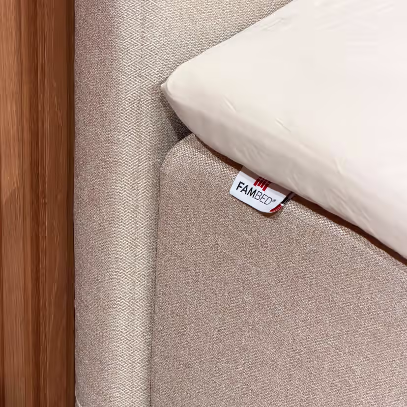 Close-up van de beige hoes rond de hoek van het bed, met een FAMBED-label en een wit laken erop
