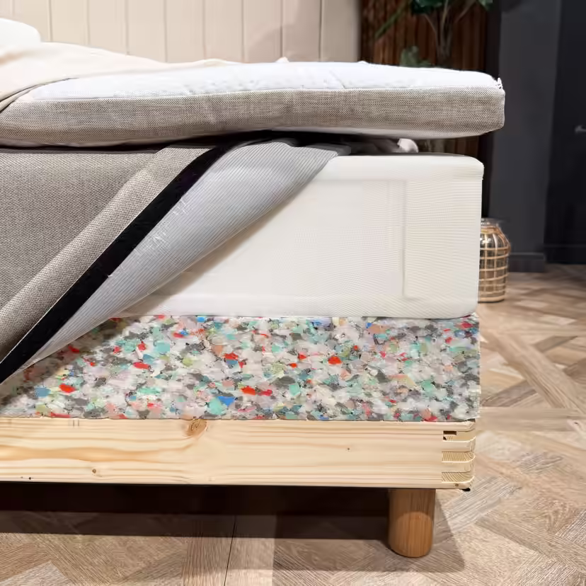 De lagen van het bed zijn bij de hoek geopend: een topmatras, een binnenveringsmatras met geopende hoes en de schuimrand van de bedbodem boven het houten frame